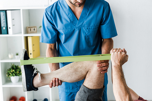 cropped view of bearded doctor holding leg of mature man exercising with elastics