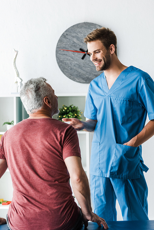 happy doctor standing with hand in pocket near mature man in clinic