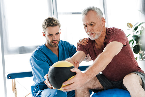 doctor sitting near mature patient exercising on fitness ball