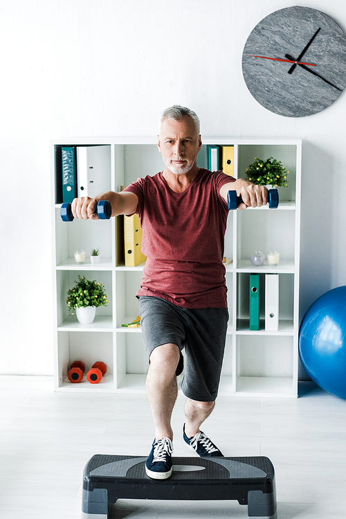 bearded middle aged man exercising with dumbbells on step platform in clinic