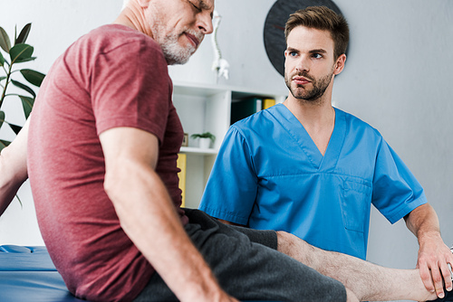 selective focus of handsome bearded doctor holding leg on mature patient with pain