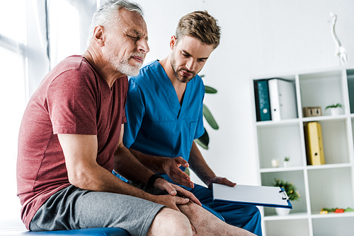 bearded mature patient with closed eyes touching leg near doctor holding clipboard in clinic