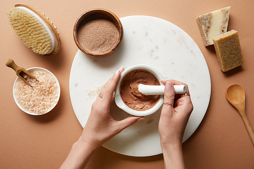 cropped view of woman mixing clay in bowl on marble circle near organic cosmetic ingredients and massage brush on beige background