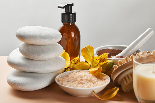 selective focus of spa cosmetics, stones, massage brush, candle and yellow flowers on beige surface isolated on grey