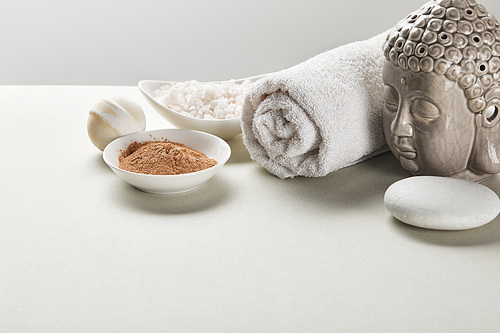 씨솔트 and clay powder in bowls, cotton towel, stone, bath bomb and buddha figurine on white table isolated on grey