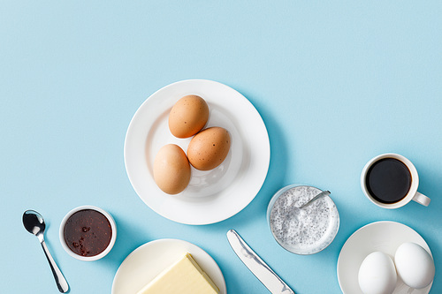 top view of fresh boiled eggs, butter, jam on white plates, yogurt with chia seeds, coffee, spoon and knife on blue background