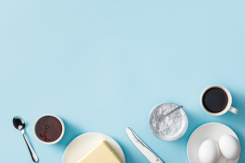 top view of butter, jam, yogurt with chia seeds, coffee and fresh eggs on white plates, knife and spoon on blue background
