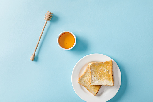 top view of honey in bowl, wooden dipper and two toasts on white plate on blue background