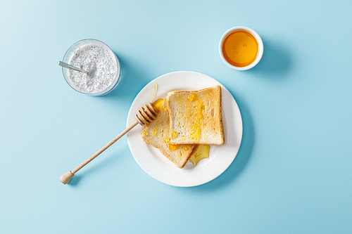 top view of yogurt with chia seeds, toasts with honey and honey dipper on white plate on blue background