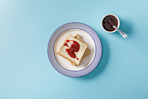 top view of toasts on plate and spoon in bowl with jam on blue background