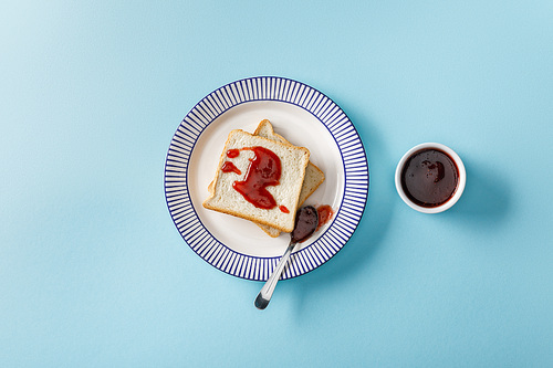 top view of toasts and spoon with jam on plate near bowl on blue background