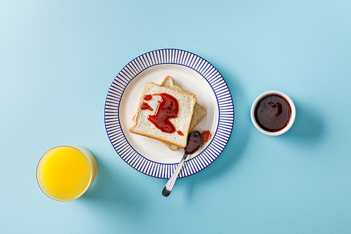 top view of orange juice, toast, bowl and spoon with jam on ornamental plate on blue background