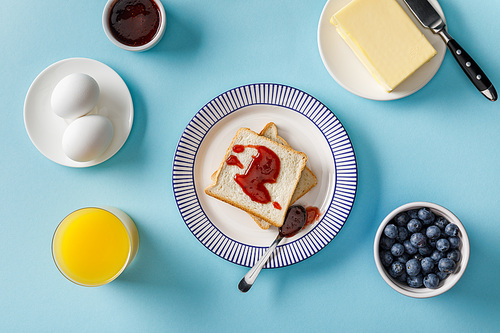 top view of toasts with jam, orange juice, boiled eggs, butter and blueberries on blue background
