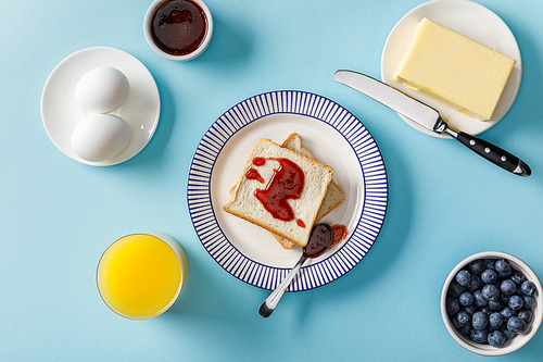 top view of toasts, bowl with jam, spoon and knife on plates, boiled eggs, butter, blueberries and orange juice on blue background