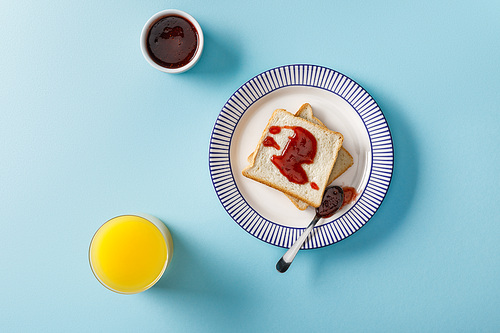 top view of orange juice, toasts, bowl and spoon with jam on plate on blue background