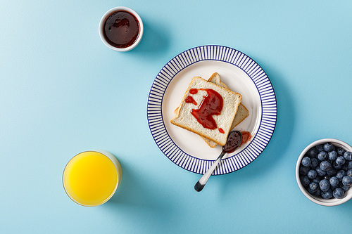 top view of orange juice, blueberries, toasts, bowl and spoon with jam on plate on blue background
