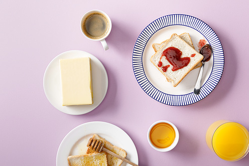 top view of butter, coffee, orange juice, toasts with honey and jam on plates on violet background