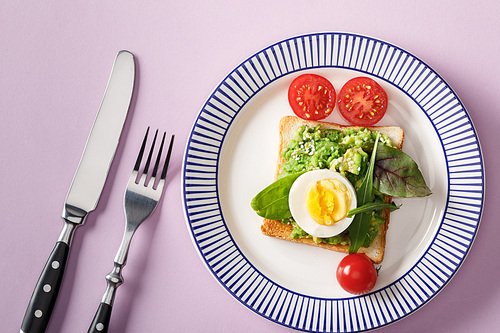 top view of toast with guacamole, boiled egg, spinach, cherry tomatoes, fork and knife on ornamental plate on violet background