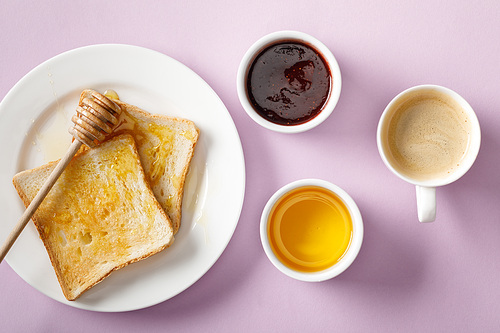 top view of bowls, toasts with honey on white plate, wooden dipper, jam and cup of coffee on violet background