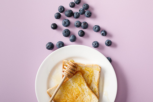 top view of toasts with honey, wooden dipper on white plate and scattered blueberries on violet background