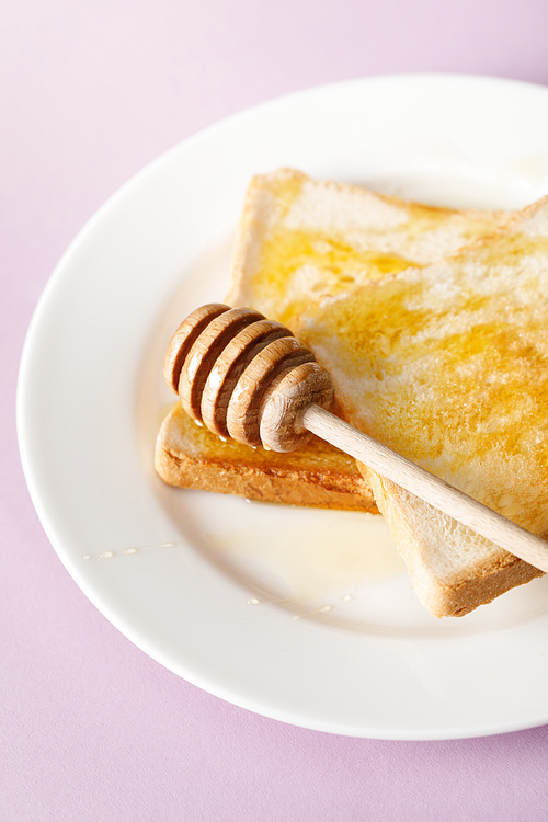 close up view of toasts with honey and wooden dipper on white plate on violet background
