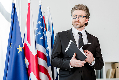 bearded diplomat holding folder near flags