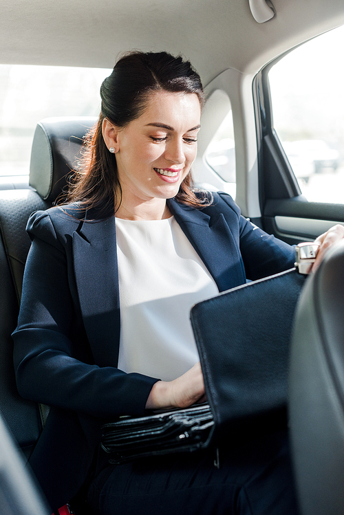 attractive ambassador smiling while sitting in car and touching briefcase