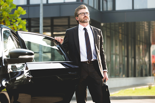 handsome diplomat in glasses standing with briefcase near black car