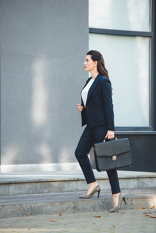 side view of attractive diplomat holding briefcase and walking near building