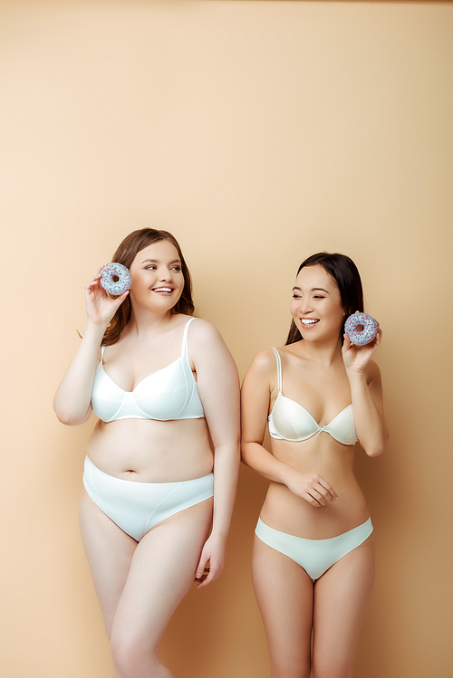 happy plus size woman and asian girl in underwear holding doughnuts isolated on beige, body positive concept