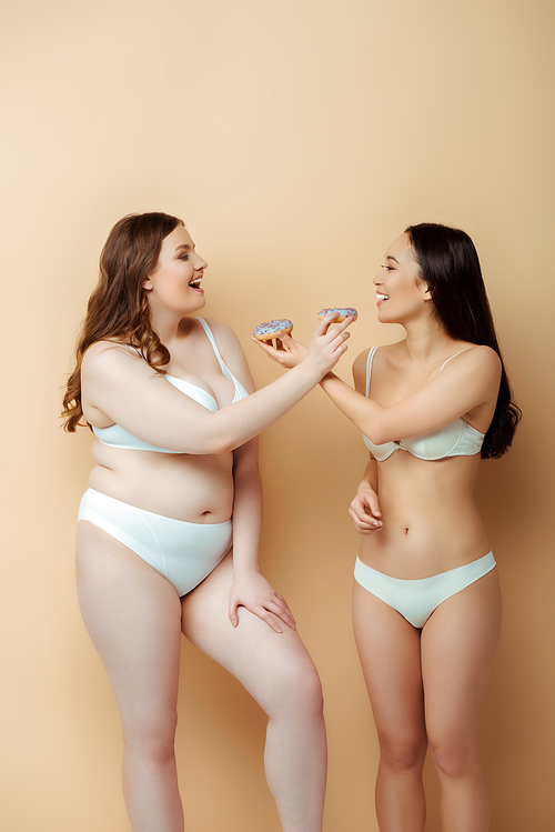 profile of happy plus size woman and asian girl in underwear holding doughnuts isolated on beige, body positive concept