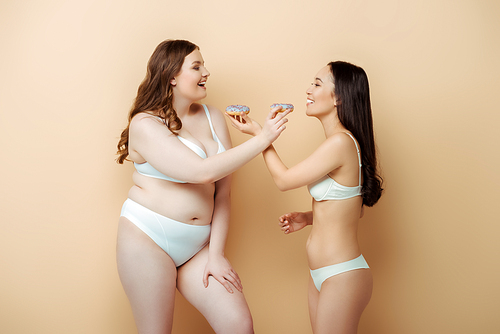 cheerful plus size woman and asian girl in underwear holding doughnuts isolated on beige, body positive concept