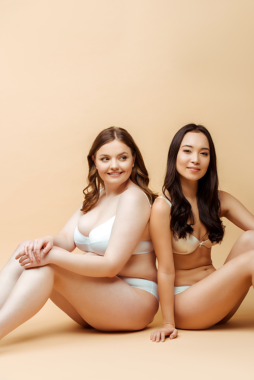 cheerful overweight woman and asian girl in underwear sitting on beige, body positive concept