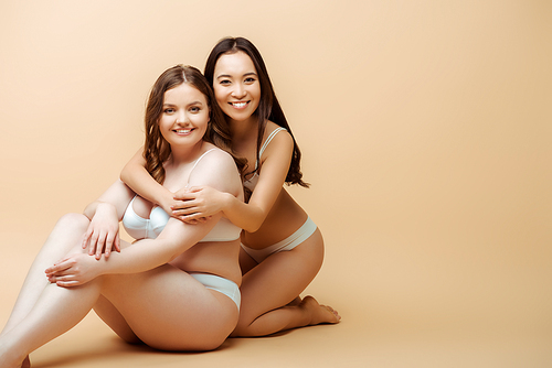 cheerful asian girl hugging overweight woman in underwear while sitting on beige, body positive concept
