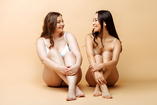 cheerful plus size woman and smiling asian girl in lingerie sitting on beige, body positive concept