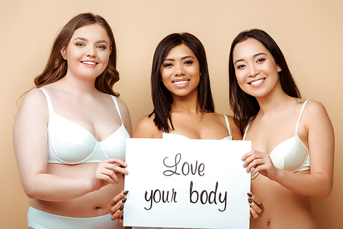 cheerful and overweight multicultural women in bras holding placard with love your body lettering isolated on beige, body positive concept