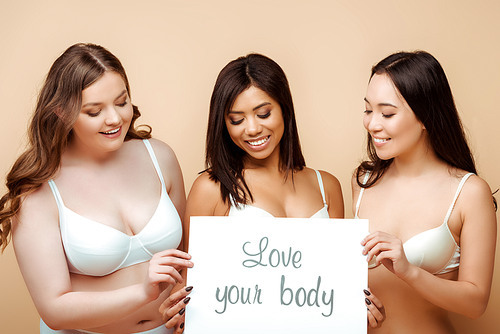 happy and overweight multicultural women in bras holding placard with love your body lettering isolated on beige, body positive concept