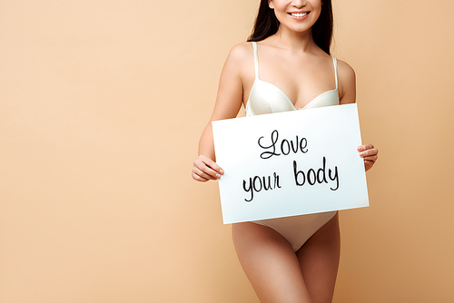 cropped view of cheerful girl holding placard with love your body lettering isolated on beige