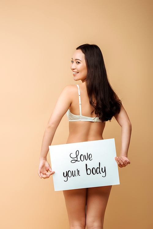 cheerful and asian woman holding placard with love your body lettering isolated on beige