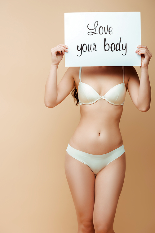 young woman covering face while holding placard with love your body lettering isolated on beige