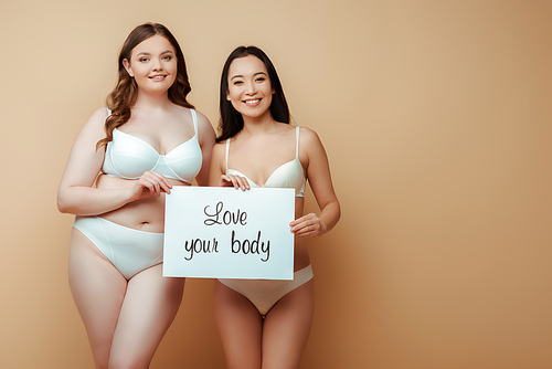 happy multicultural girls in underwear holding placard with love your body lettering isolated on beige, body positive concept