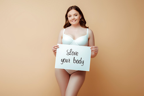 happy plus size girl holding placard with love your body lettering isolated on beige, body positive concept