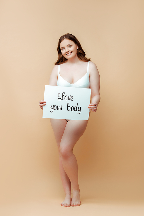 Plus size woman smiling,  and holding placard with love your body inscription on beige