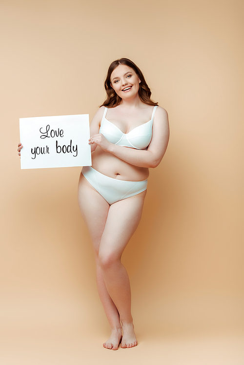 Plus size woman smiling,  and showing placard with love your body lettering on beige