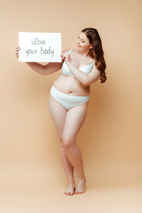 Plus size woman smiling and holding placard with love your body inscription on beige background