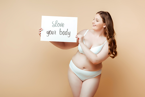 Plus size woman smiling and showing placard with love your body lettering on beige