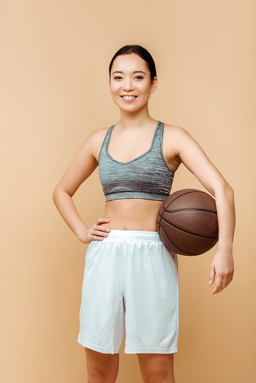 Asian sportswoman with hand on hip and ball smiling and  isolated on beige