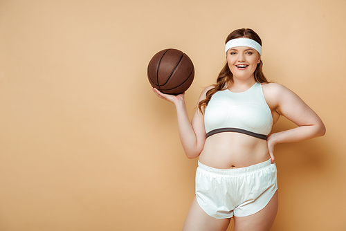 Plus size sportswoman with hand on hip and ball smiling and  on beige