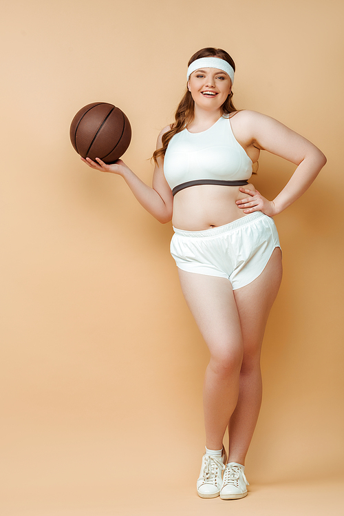 Plus size sportswoman with hand on hip smiling, holding ball and  on beige background