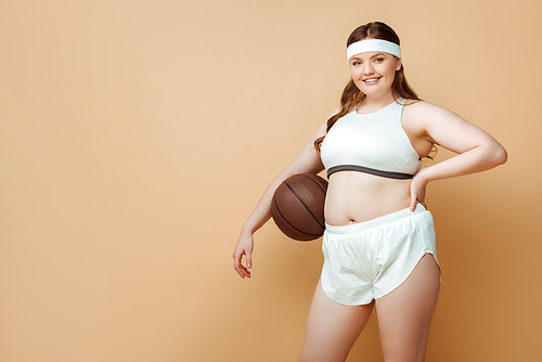 Plus size sportswoman with ball and hand on hip smiling and  on beige